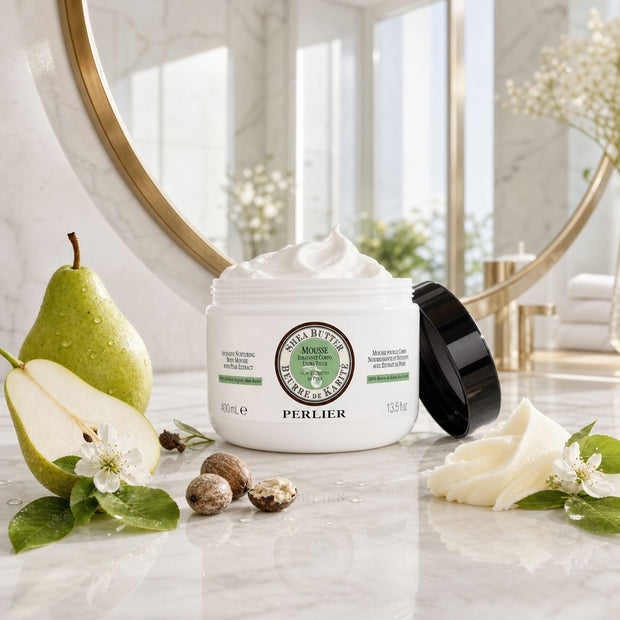 13.5 oz jar of Perlier Shea Pear Body Mousse on a marble surface with pears and flowers, large round mirror in the background