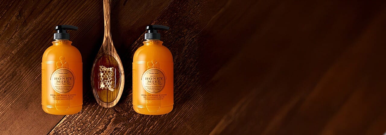 Two bottles of Perlier Honey Miel Bath & Body Wash with a wooden spoonful of honey on a dark wooden background