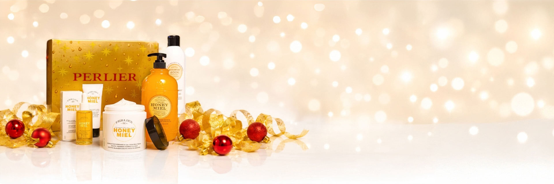 A Perlier holiday gift set featuring a gold glittery box, and assorted Honey Miel products, styled with gold ribbons and red ornaments on a white surface against a warm bokeh background.