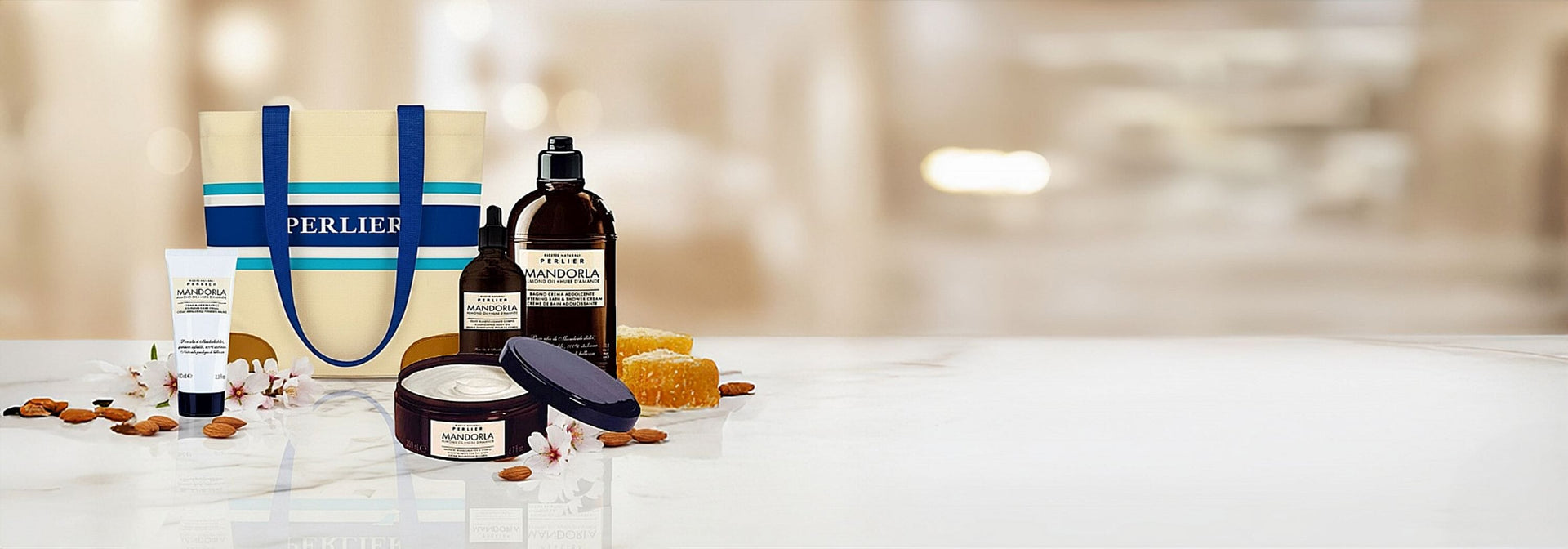 Perlier 4 Mandorla Almond bath and body products and a tote bag on a white surface next to almonds and honey, with a blurred background.