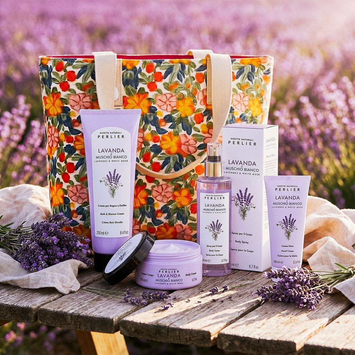 4 Perlier Lavender & White Musk bodycare products and a floral tote bag on a wooden table next to a lavender field.