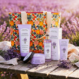 4 Perlier Lavender & White Musk bodycare products and a floral tote bag on a wooden table next to a lavender field.