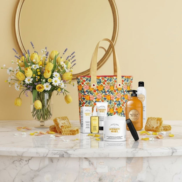 5 Perlier Honey Miel body care products and a floral tote bag, in front of a floral arrangement on a marble surface with a decorative mirror in the background.