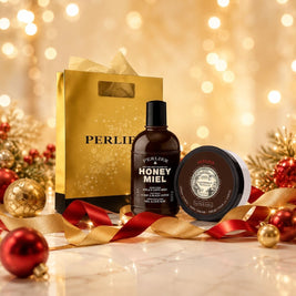 A gift set featuring a Perlier Honey Miel Coffe body wash and a Caribbean Vanilla Coffe body cream, and a gold gift bag styled with gold and red Christmas ornaments and ribbons on a marble surface with a warm bokeh background.