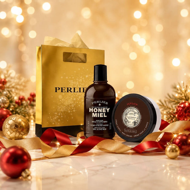 A gift set featuring a Perlier Honey Miel Coffe body wash and a Caribbean Vanilla Coffe body cream, and a gold gift bag styled with gold and red Christmas ornaments and ribbons on a marble surface with a warm bokeh background.