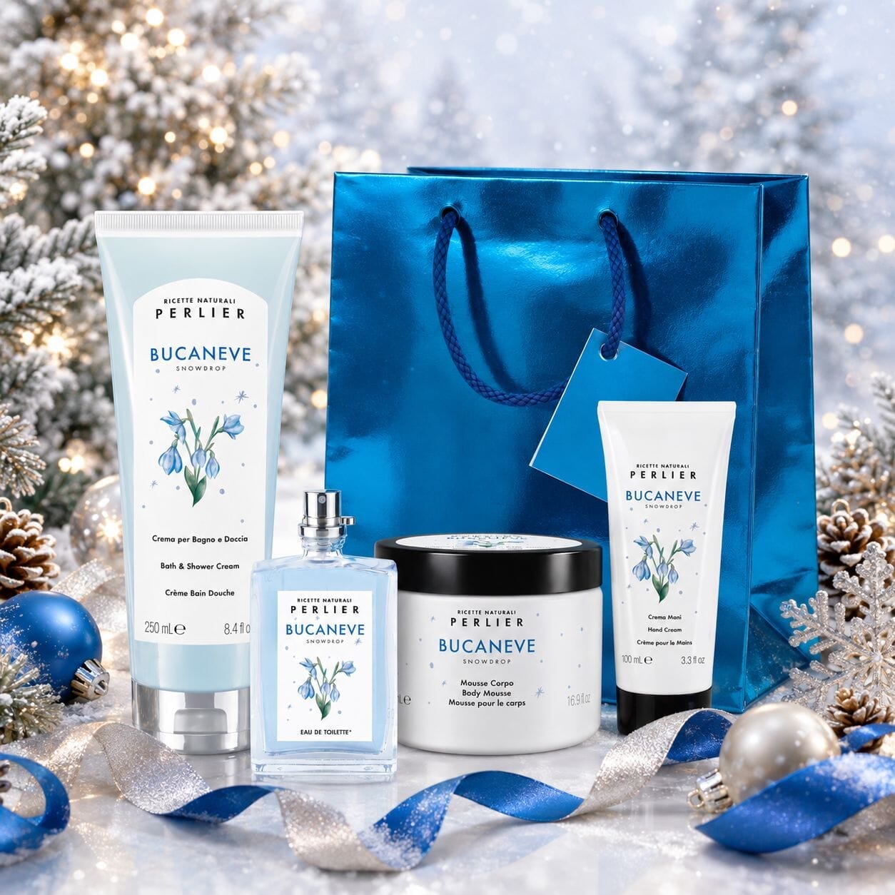A Perlièr Bucaneve gift set arranged on a snowy festive surface with pine branches, ornaments, and ribbons. The set includes Perlier Snowdrop bath & shower cream, body mousse, hand cream and perfume with a metallic blue gift bag.