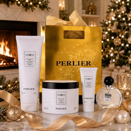 A holiday gift set from Perlier featuring The Musk products including a body wash, hand cream, body cream, and perfume arranged with a large gold Perlier shopping bag, and festive holiday decorations in front of a cozy fireplace.