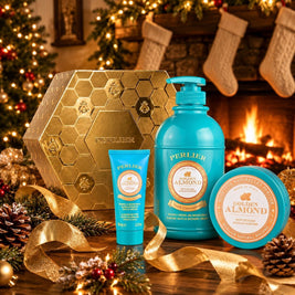 A Perlier Golden Almond gift set featuring a body wash, body balm, and hand cream, arranged on a table with gold ribbons, pinecones, and a warm fireplace background.