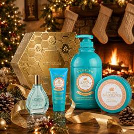 A Perlier Golden Almond gift set featuring a body wash, body balm, hand cream, and perfum, arranged on a table with gold ribbons, pinecones, and a warm fireplace background.