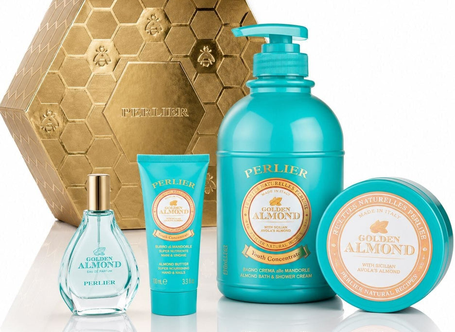 A Perlier Golden Almond gift set featuring a body wash, body balm, hand cream, and perfum, arranged in front of a gold hexagonal box on a white reflective surface.